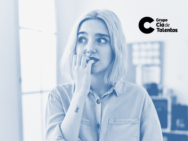 A imagem apresenta uma jovem de cabelos claros, lisos e na altura dos ombros. Ela está com uma das mãos na boca, como se estivesse roendo as unhas, demonstrando apreensão, em referência ao tema do artigo que é sobre enfrentar a timidez em processos seletivos.