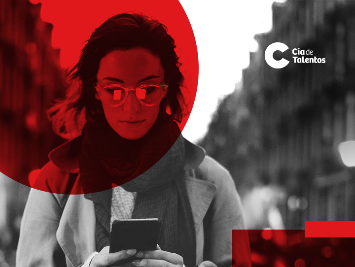 A imagem que ilustra o artigo Vaga em tecnologia: como conseguir uma traz a foto de uma mulher em preto e branco. Sobre o rosto dela há um círculo vermelho. Ela é branca, tem os cabelos lisos, usa óculos, está com um celular nas mãos olhando para o aparelho.