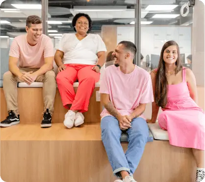 Quatro pessoas sentadas em um ambiente de escritório. Elas estão sorrindo e parecem estar conversando de maneira descontraída. Os rapazes vestem rosa e se olham, já as moças olham para frente e vestem vermelho e rosa.