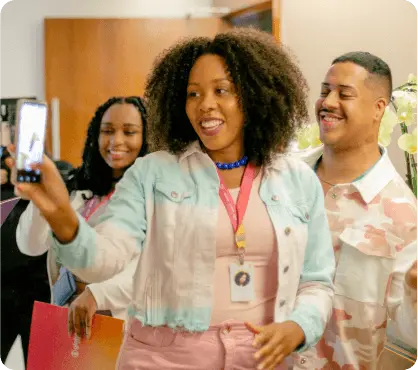 Um grupo de três pessoas em um ambiente interno, com uma mulher negra em destaque segurando um celular e tirando uma selfie. Todos parecem estar felizes e engajados na atividade.