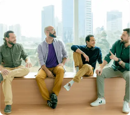 Quatro homens sentados diante de uma grande janela que mostra uma paisagem urbana ao fundo. Eles estão conversando entre si e aparentam estar em um momento de descontração.